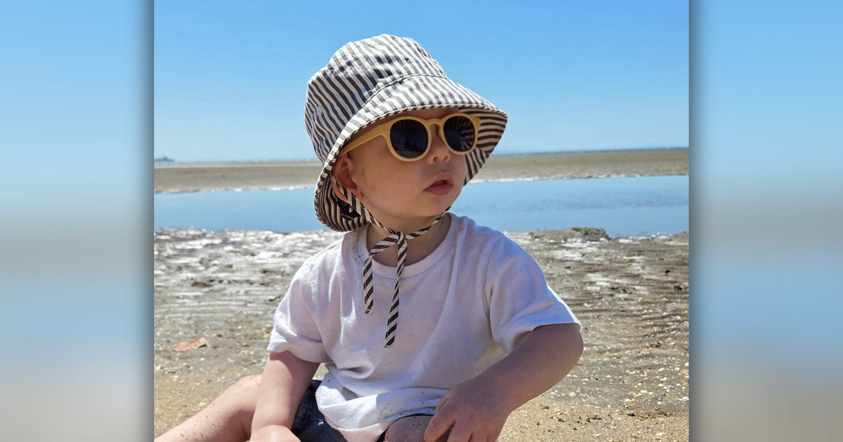 Toddler at the beach wearing a striped bucket hat and peach Hey Peeps Little Keypers sunglasses.